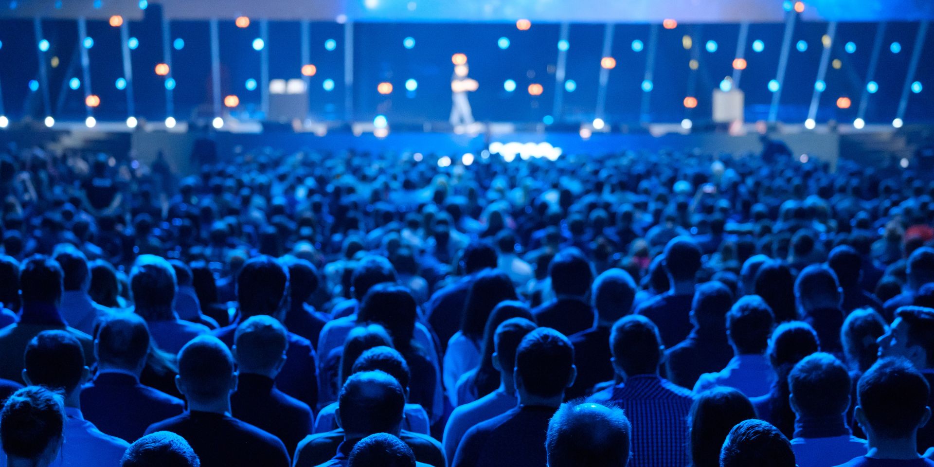 A blue-lit room of conference visitors listen to a talk on stage.