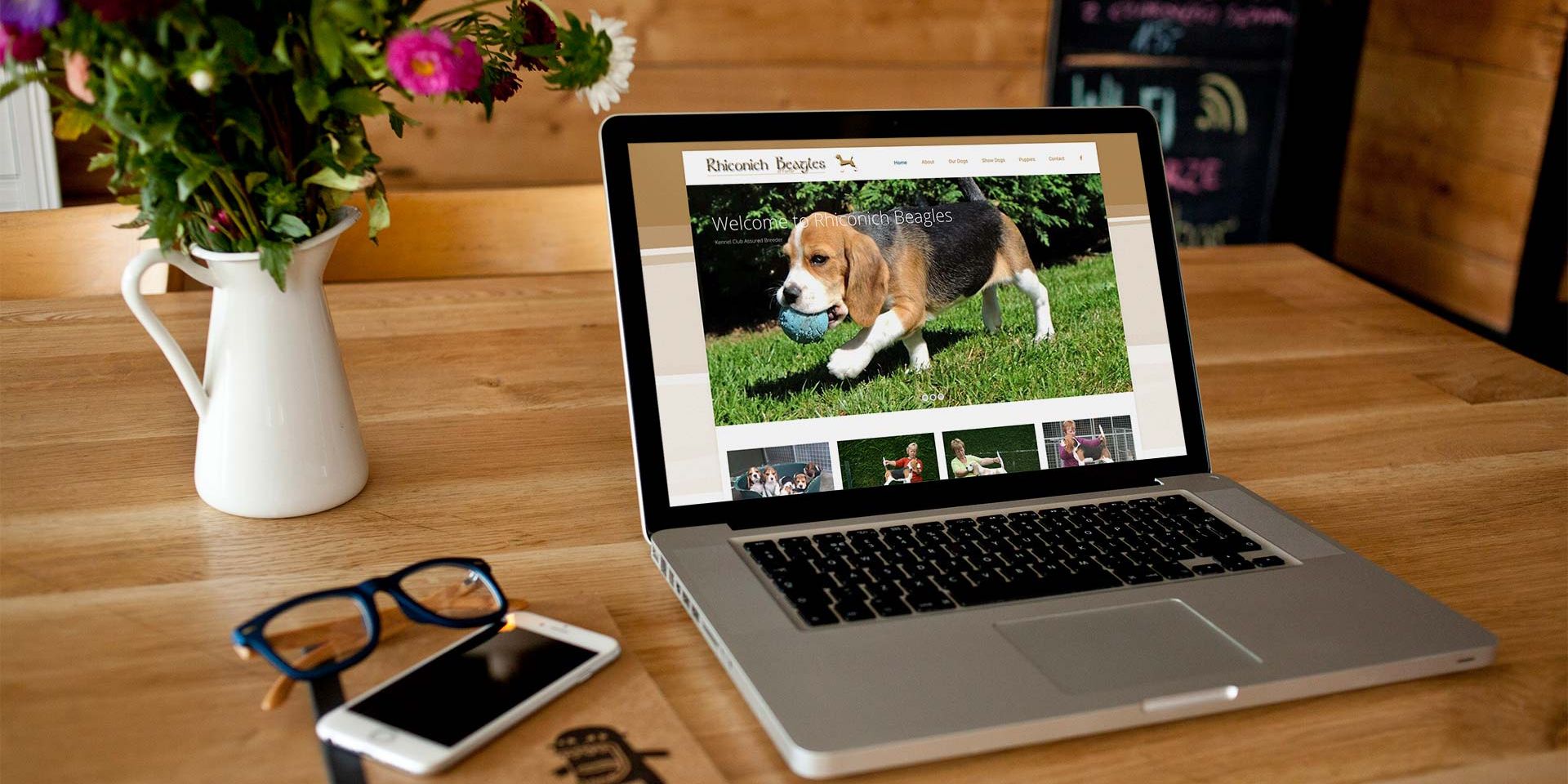 This image shows a laptop placed on a wooden table beside a vase of flowers and glasses. The laptop shows a beagle dog standing on grass.