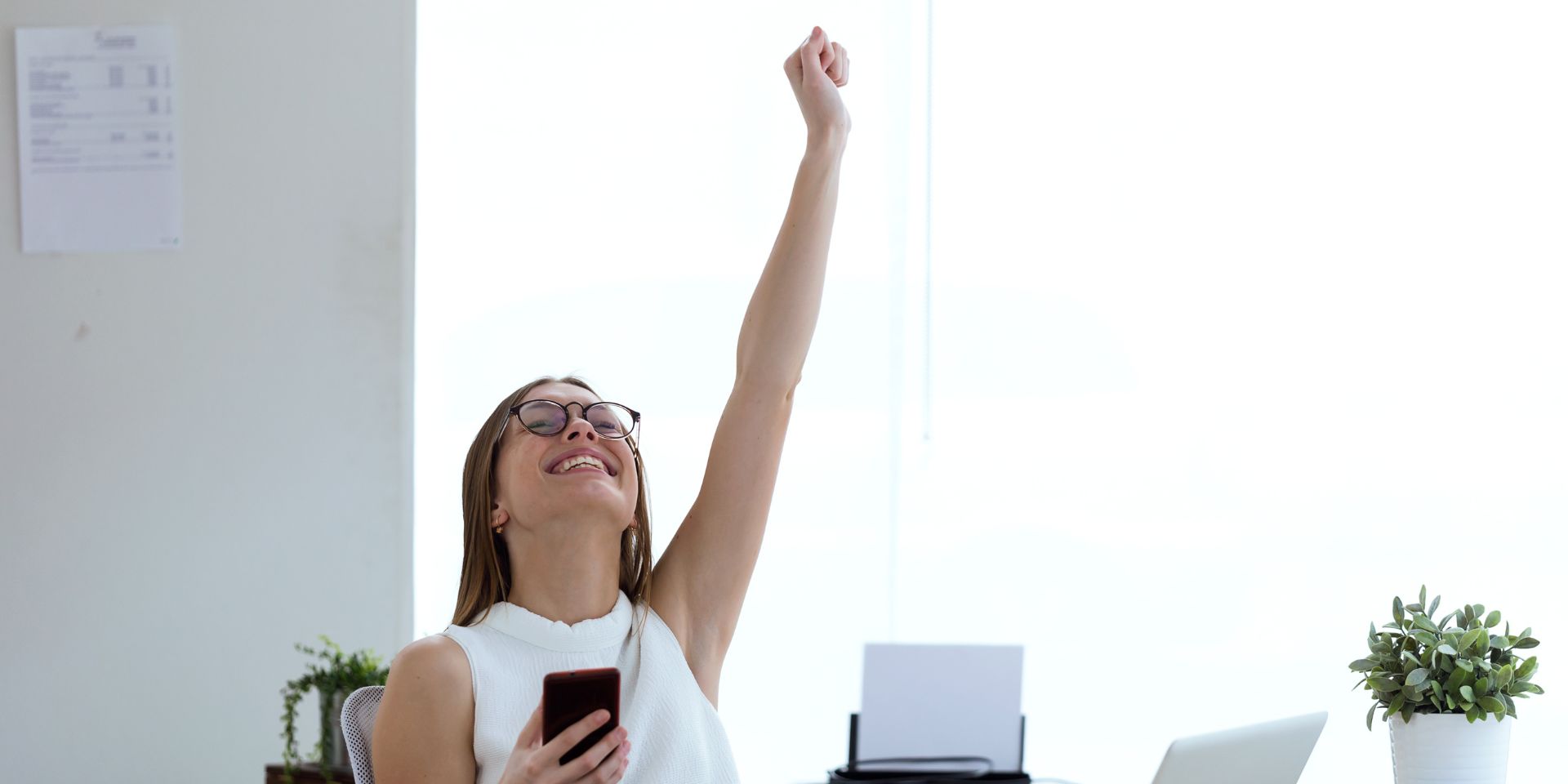 A female business professional sits at her desk celebrating with her arm in the air.