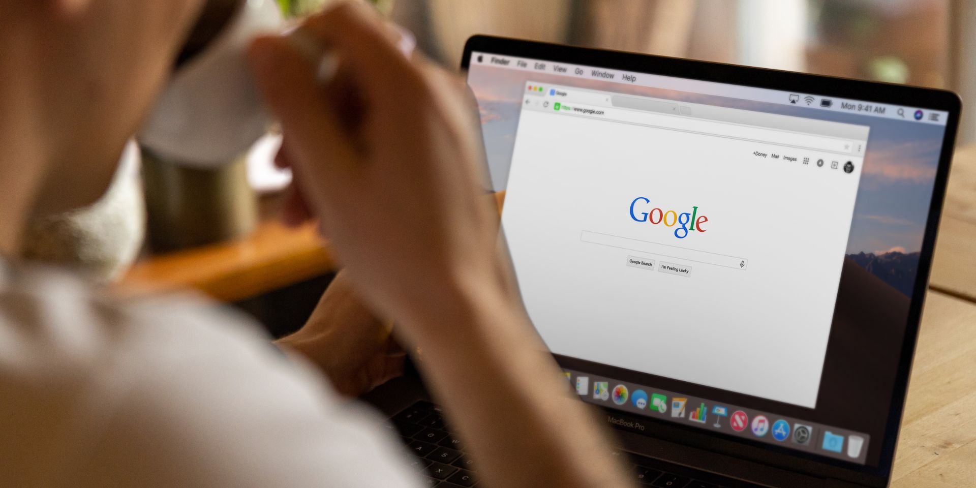 A laptop screen showing the Google website centred, with a person drinking from a cream coffee cup in the foreground.