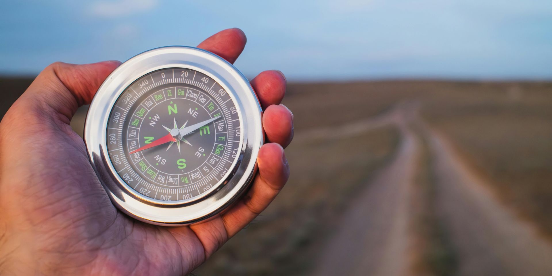A man's hand holding a silver bezel compass towards a deserted road in the background.