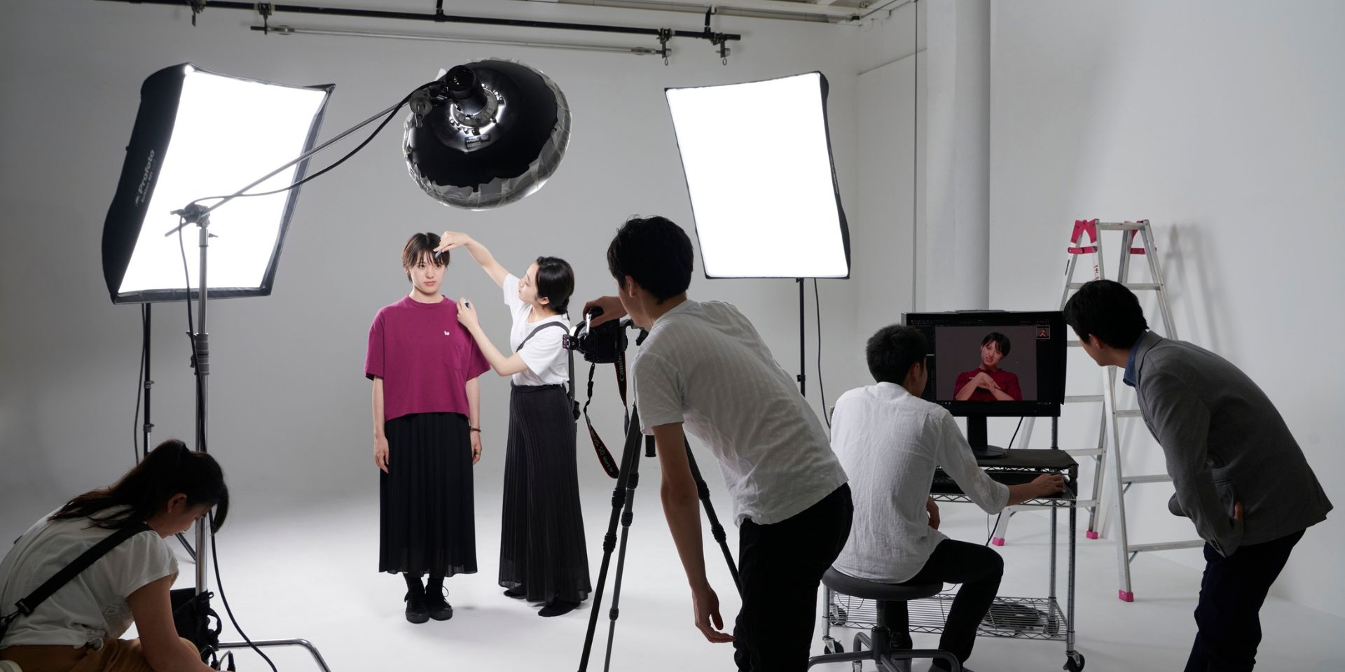A white background film set featuring a model wearing a black skirt and pink blouse, being filmed by a videography crew. Lighting and filming rigs are in place.