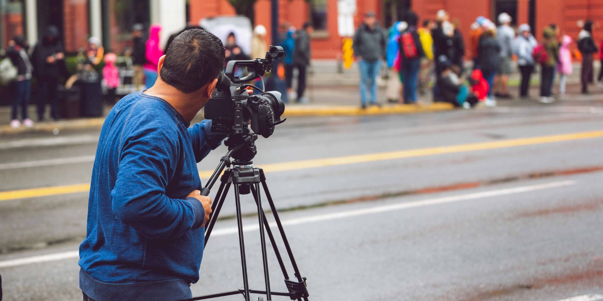 A man wearing a blue jumper, is shown to the left-side of the image filming a street scene across an empty road.