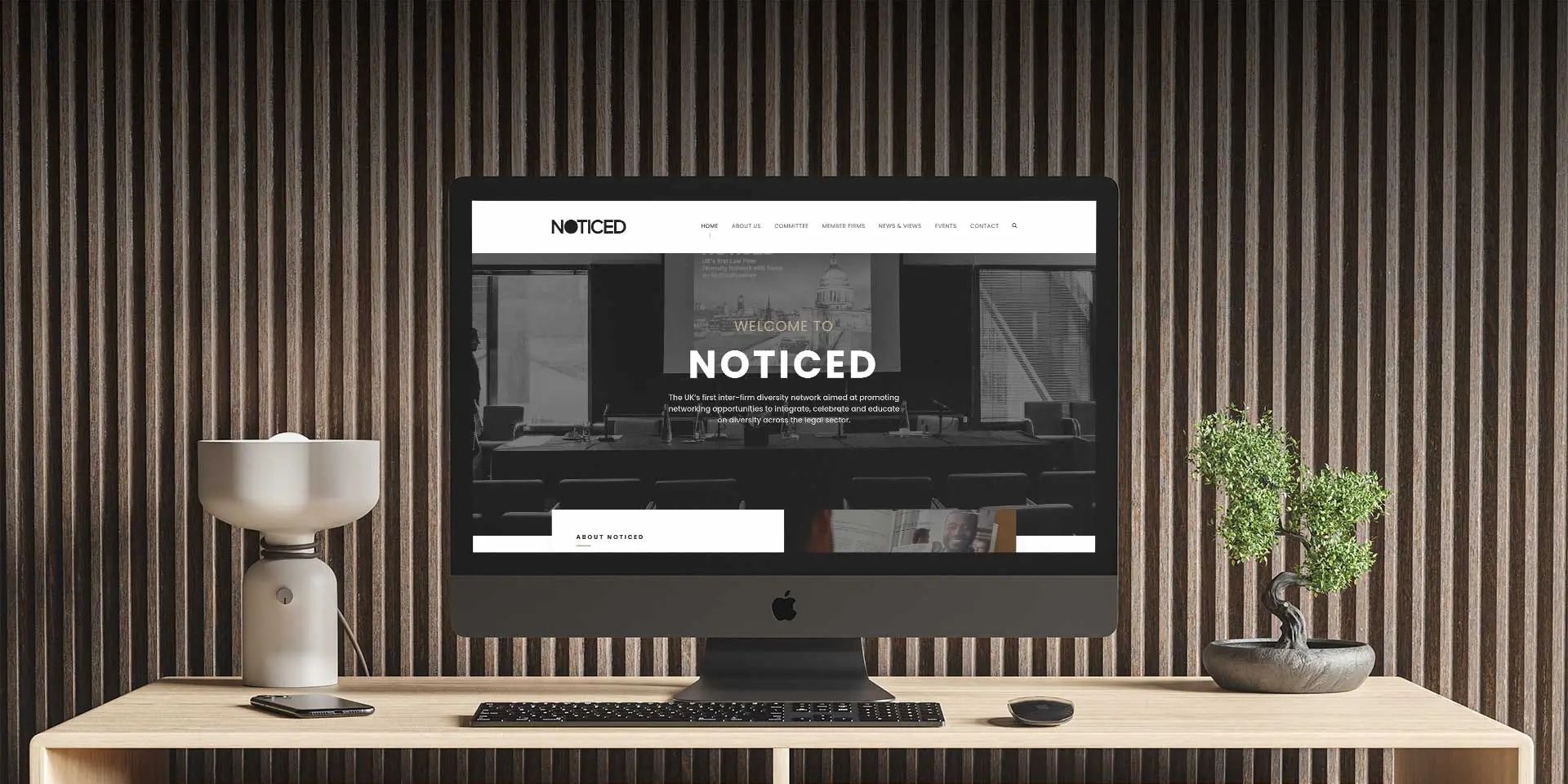 A black computer monitor with a website design features the brand name "Noticed". The background is a slatted pale wooden wall.