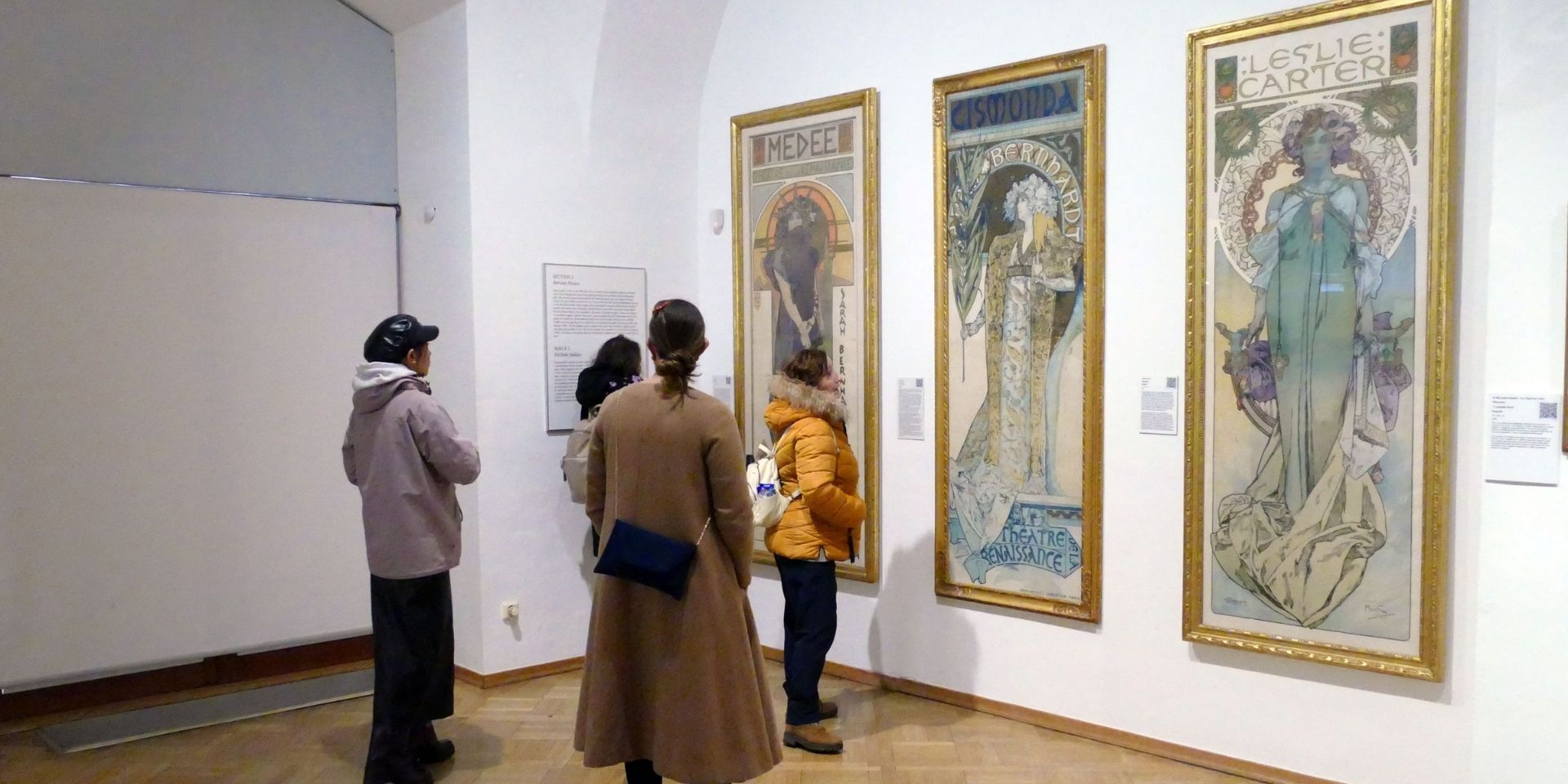 Four people stand with their backs to the camera while looking at framed artworks in a gallery setting.